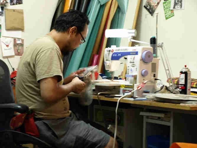 Rear, right side view of a person, sitting in front of a table with a sewing machine on it, cutting fabric with scissors