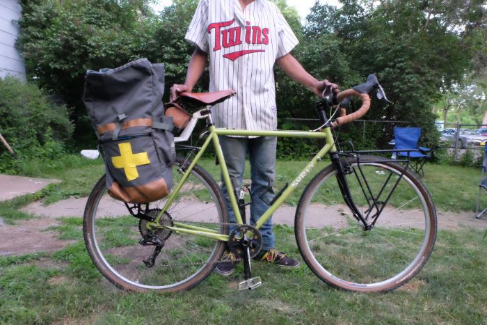 A cyclist wear a Minnesota Twins jersey stands in a front yard with a green Surly bike with a gear bag behind the seat