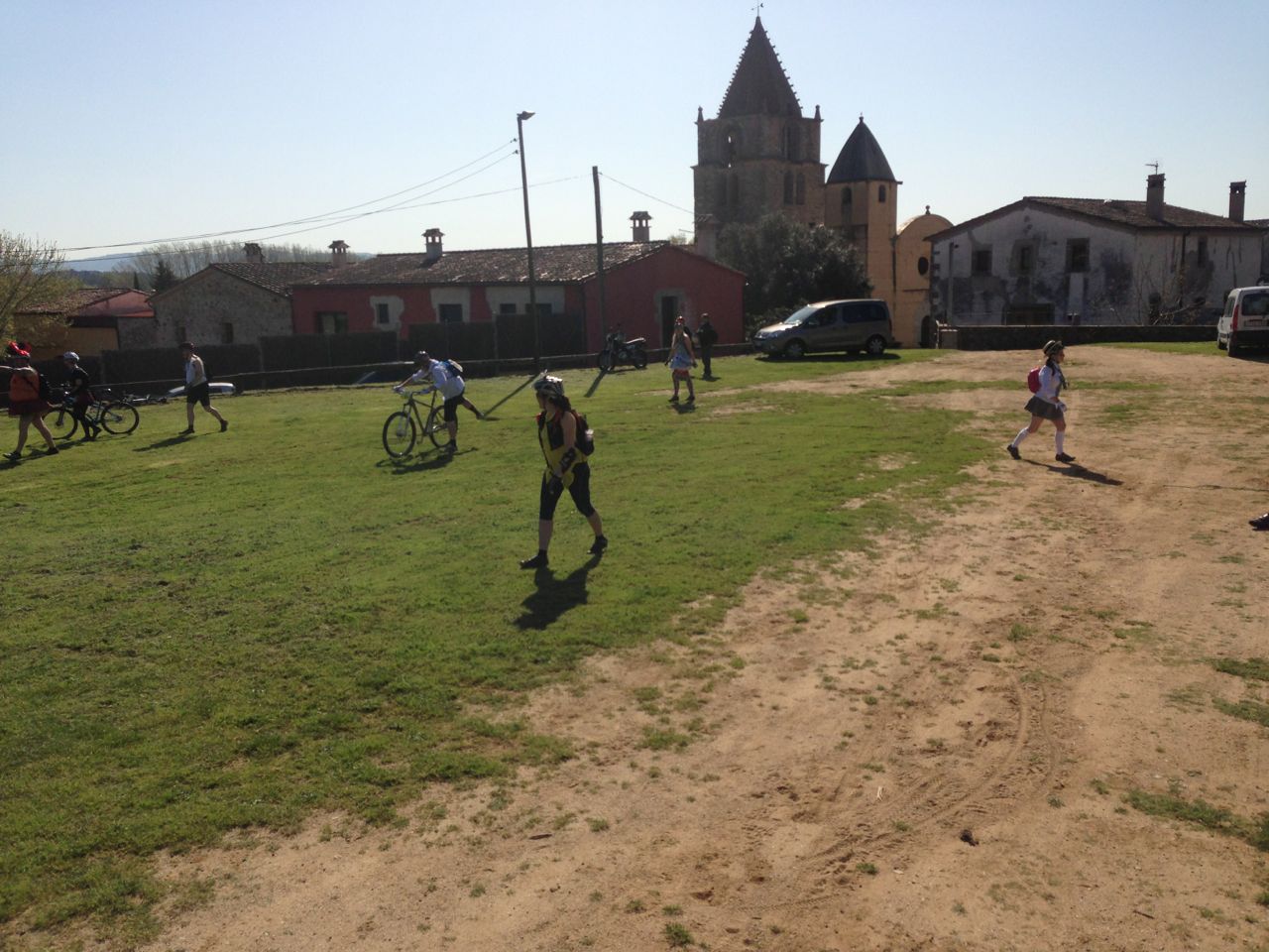 A few people walking and riding bikes, on a grass and dirt field, with building and a church behind