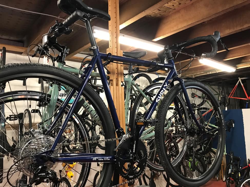 Left profile view of a blue Surly bike on rack with several bikes in the background of a room with a wood joists above