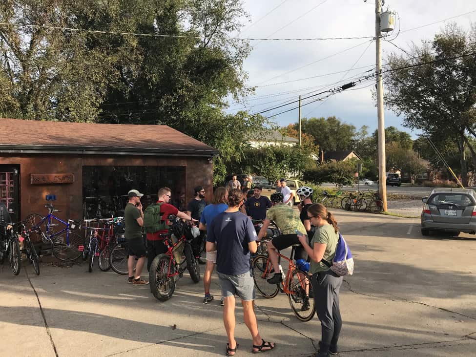 Group of cyclists standing with their bikes on a lot outside of a bike shop, with trees and power lines above