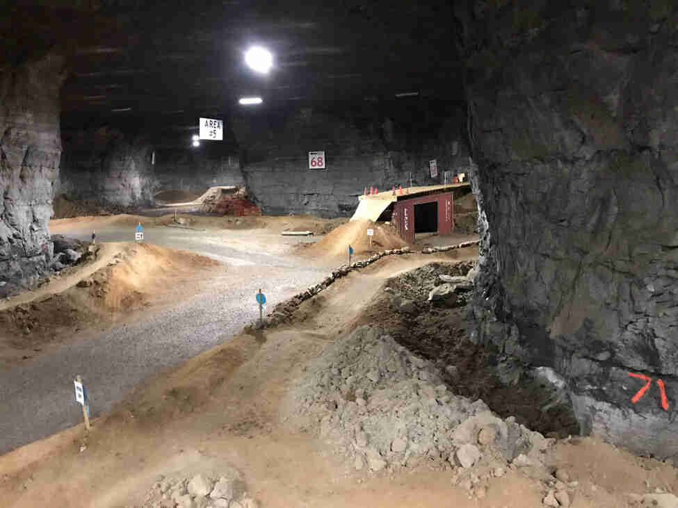 A dirt and gravel bike course with bridge structure obstacles, inside of a large cavern with lights above