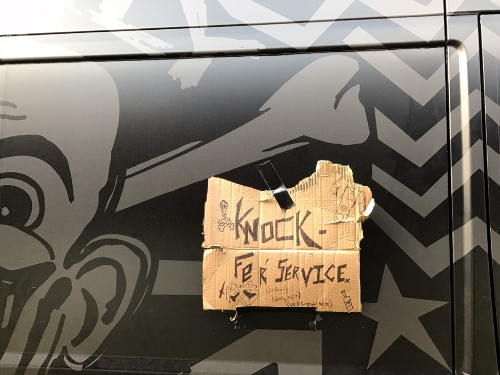 A cardboard sign with 'Knock Fer' Service' taped to the side of a black Surly van with graphics
