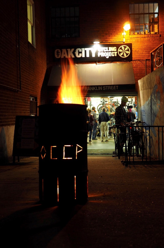 A burn barrel with a fire sits on pavement in front of the Oak City Cycling Project shop with people standing inside