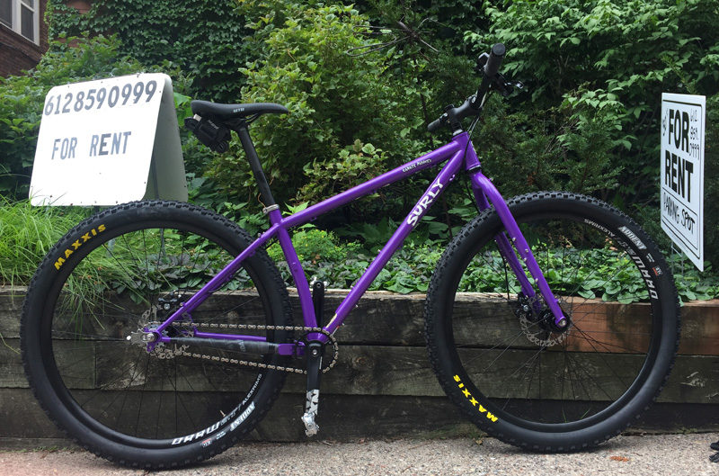 Right profile view of a Surly Karate Monkey bike, purple, leaning on a wall, next to 2 signs and green weeds behind