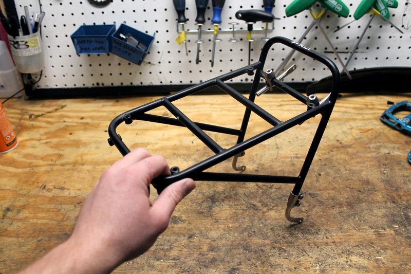 A hand holding a Surly 8-Pack rack on a wooden work bench with pegboard and tools in background