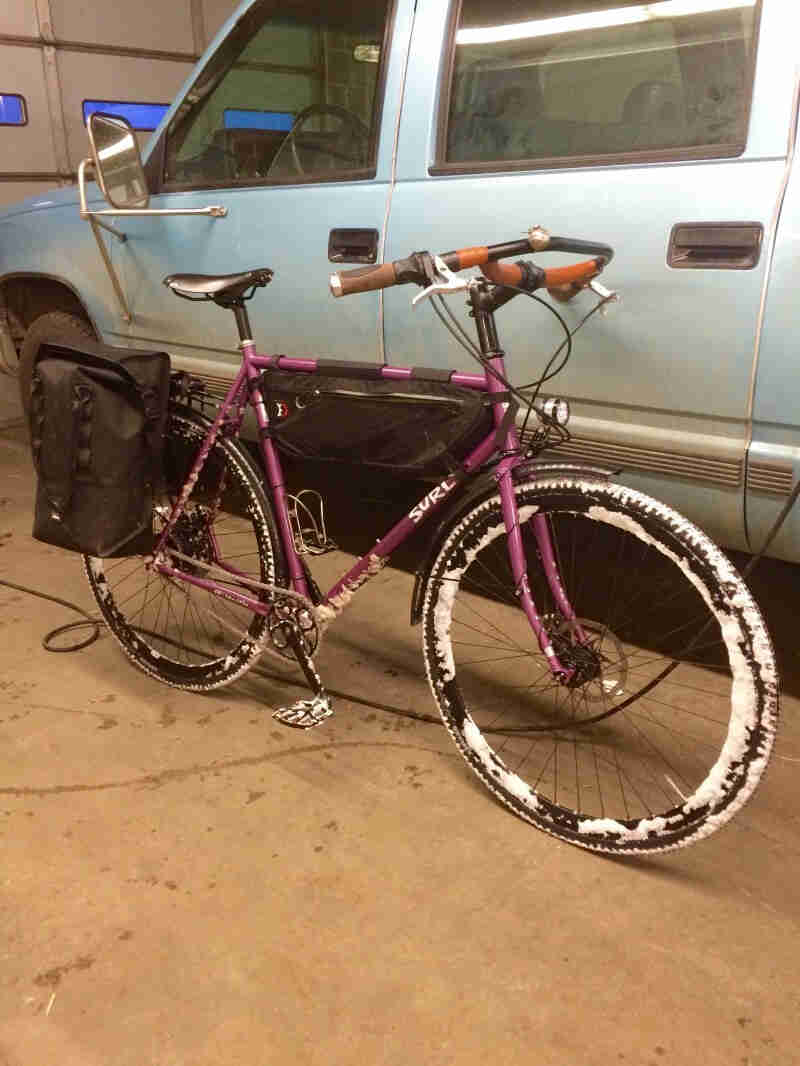 Right side view of a purple Surly Straggler bike, parked against the left side of a light blue truck, inside a garage