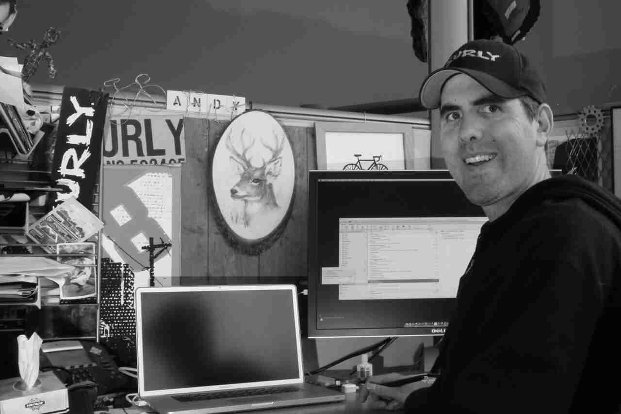 Front view of a person wearing a Surly hat, sitting at a office cubicle desk - black & white image