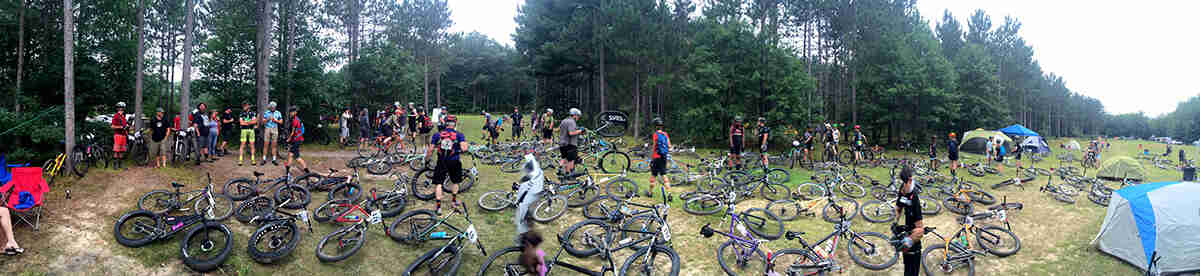 Panoramic view of a field of bikes laying on their side with wandering cyclists, and trees in the background