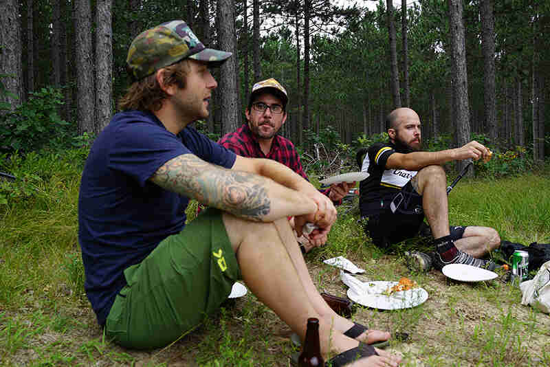 3 people sitting in the grass eating, with a pine forest in the background