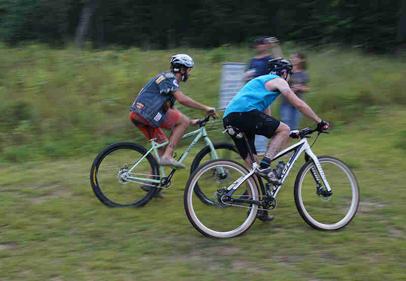 Right side view of 2 cyclists riding racing their bikes, side by side, on a grass field with trees in the background