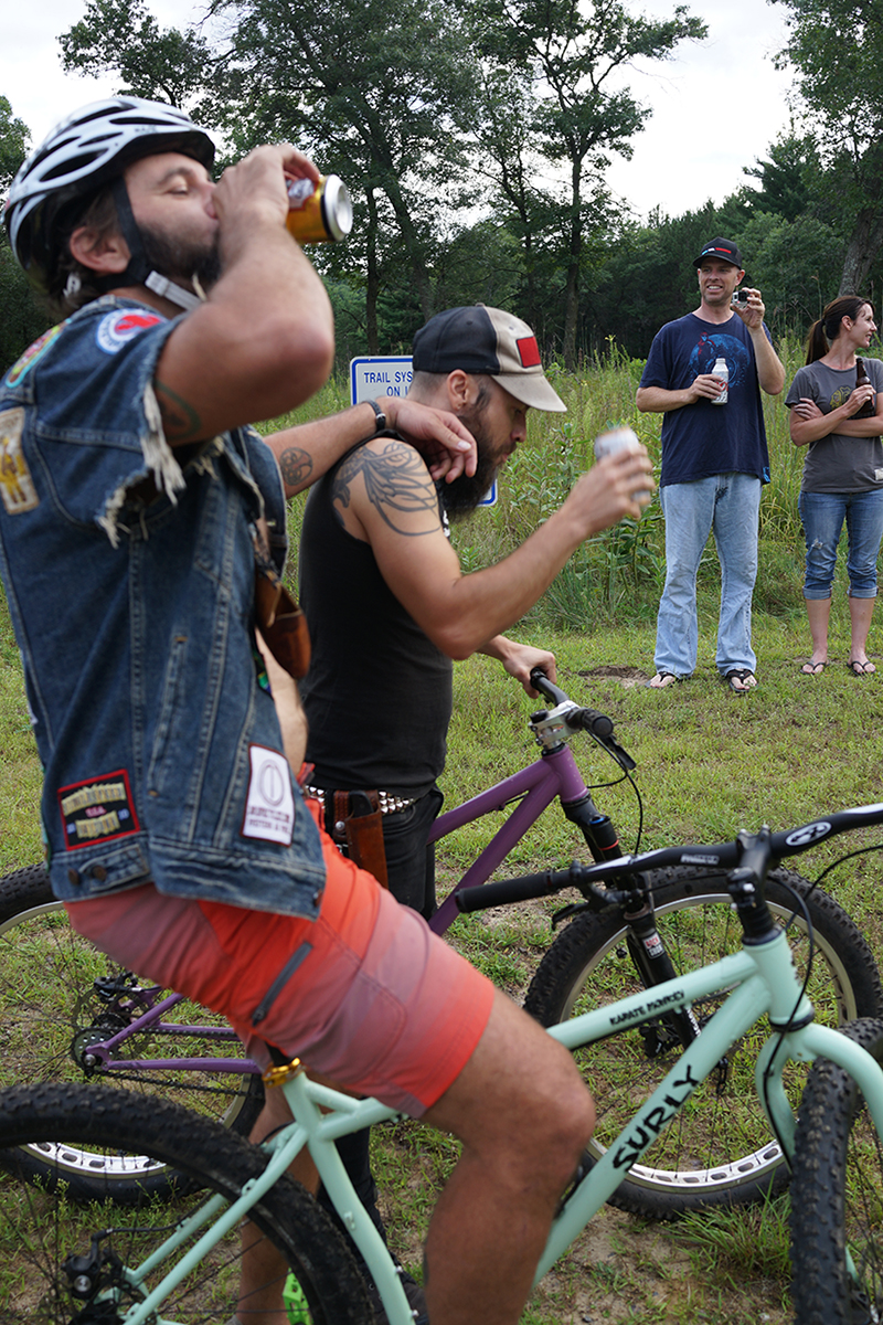 Right side view of 2 cyclist drinking beer, side by side, while standing over their Surly Karate Monkey bikes