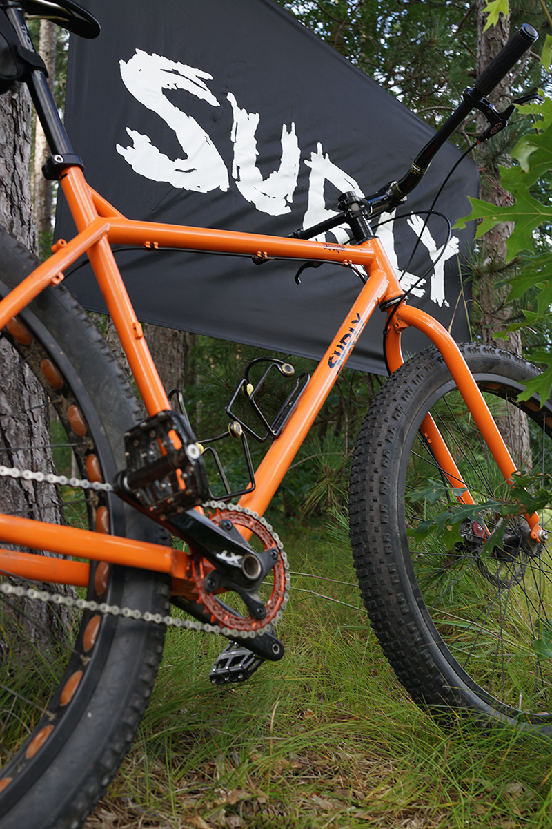 Right side view of an orange Surly Krampus XXL bike, with a Surly banner behind, with the woods in the background