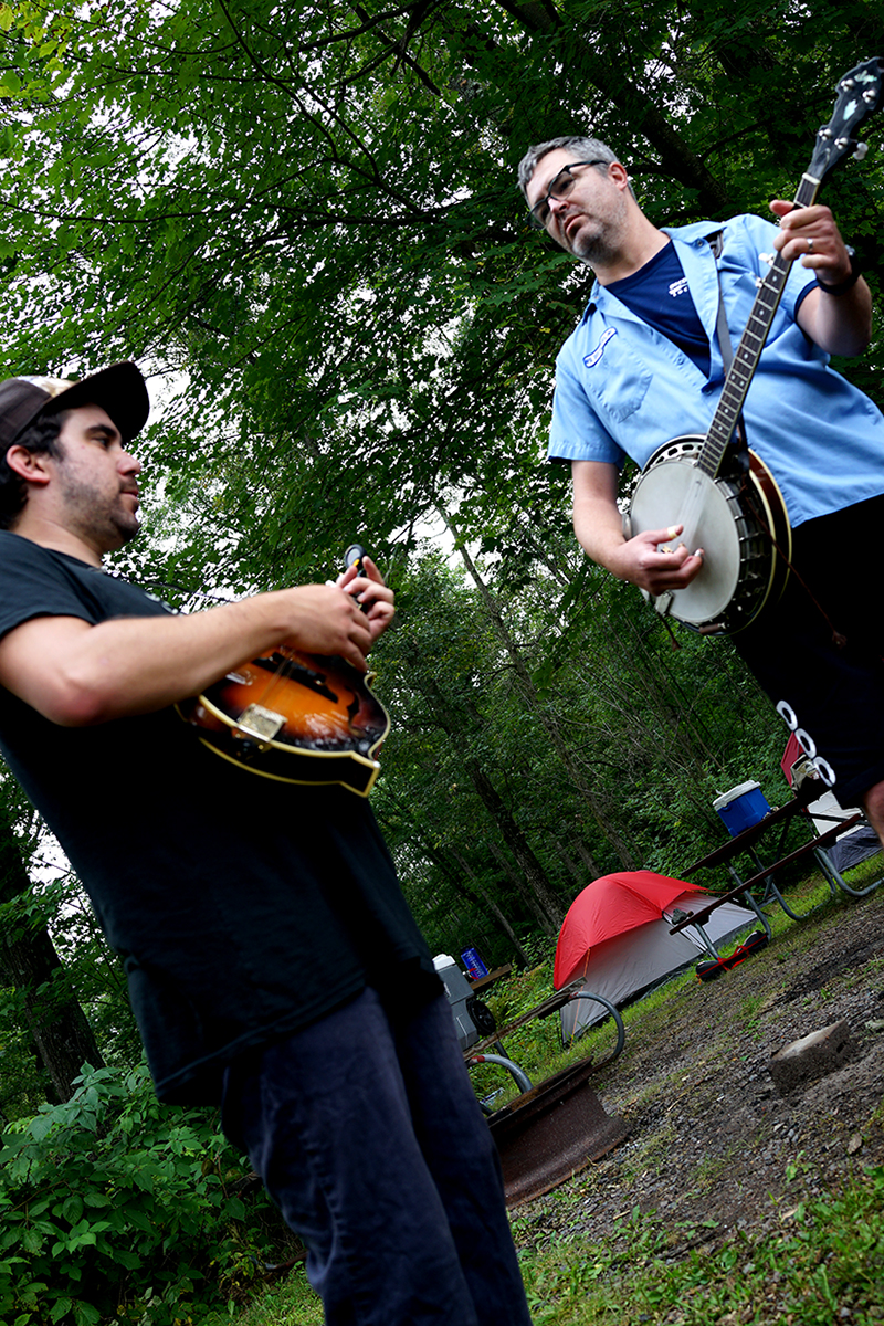 Upward, tilted view of one person playing a guitar, and another playing a banjo, at a campsite in the woods