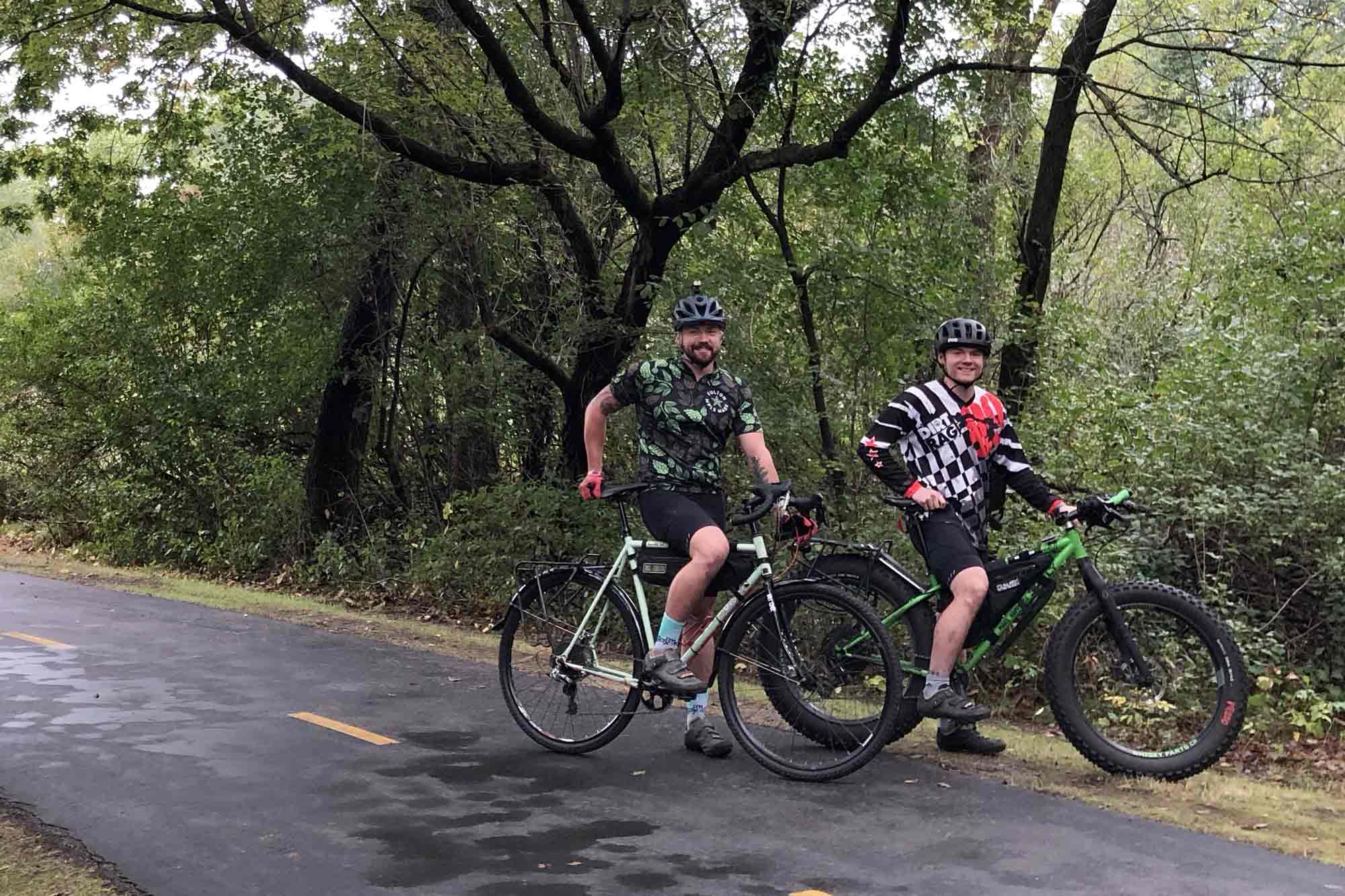 Two cyclists stand on a paved trail with their Surly bikes at a right profile view, green leafed trees in woods behind