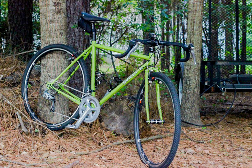 Right side view of a lime green Surly Cross Check bike, parked on pine needles in front of a tree in the woods