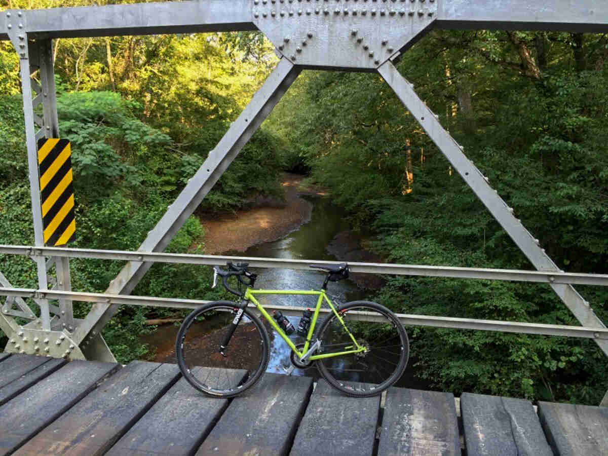 Left side view of a lime green, Surly Cross Check bike, parked on a bridge, with a stream below, in the green woods