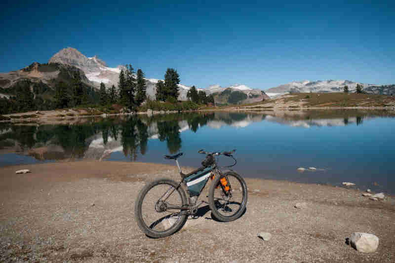 Rear, right side view of a bike on a shore in front of a lake, with trees and mountains in the background