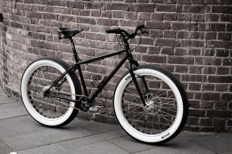 Right side view of a black Surly 1x1 bike with white wall tires, leaning against a brick wall on a sidewalk