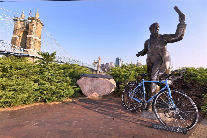Right side view of blue Surly bike, on a brick plot, in front of a bronze monument, with a bridge in the background