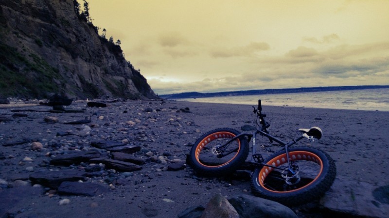 Rear view of a black Surly fat bike laying on it's left side, on a rocky sand shore, with a cliff and water ahead