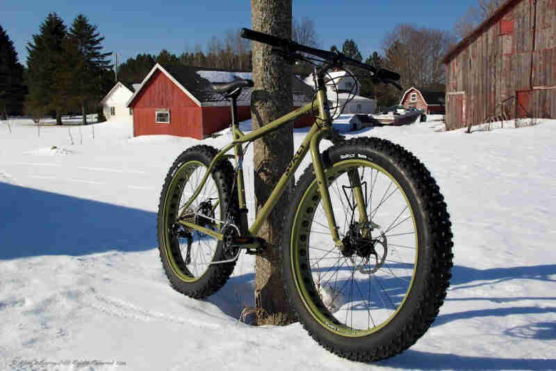 Front right side view of a green Surly fat bike, leaning on tree in a snowy field, with a farmstead in the background