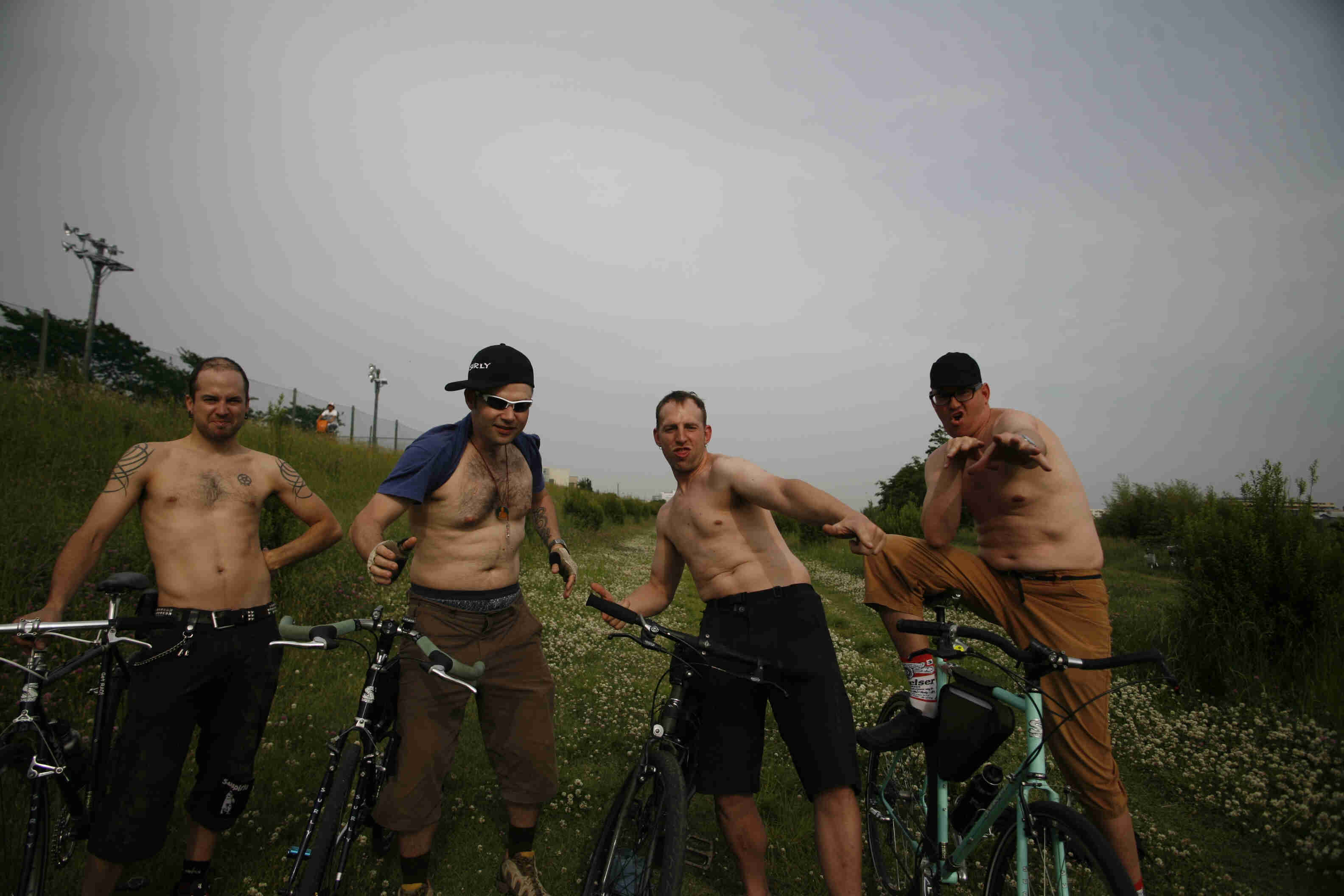 Front view of 4 people with their Surly bikes, standing side by side with no shirts, at the bottom of a grassy hill