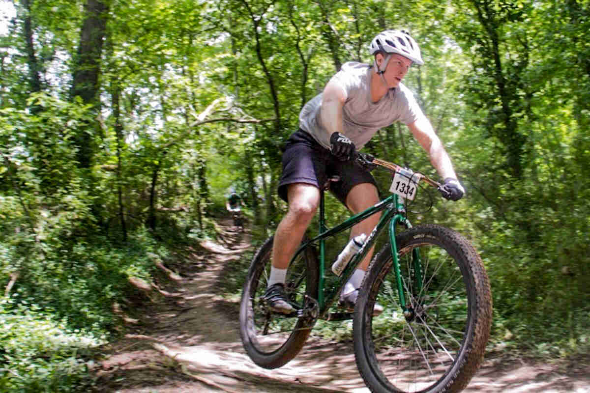 Front, right side view of a cyclist on green Surly Krampus bike, riding down a dirt trail in the woods, during a race
