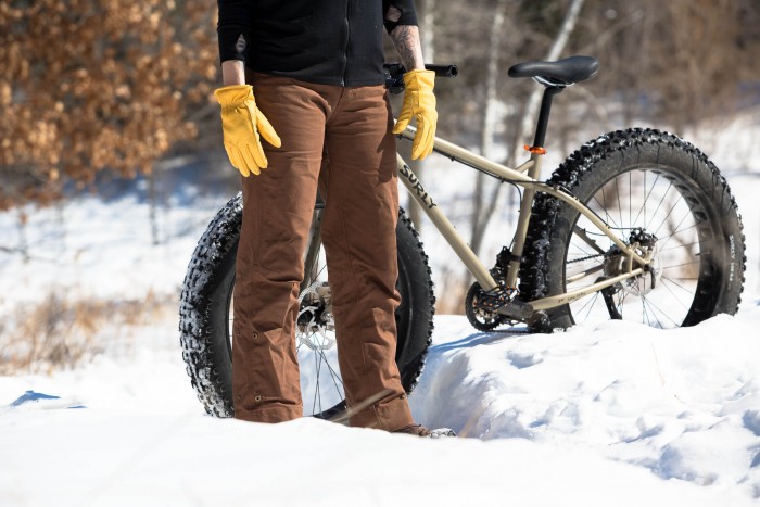 Front, waist down view of a person wearing Surly biking pants, standing in snow, with a fat bike behind them