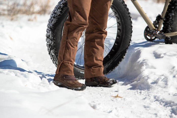 Front, thigh down view of a person wearing Surly biking pants, standing in snow, with a fat bike behind them