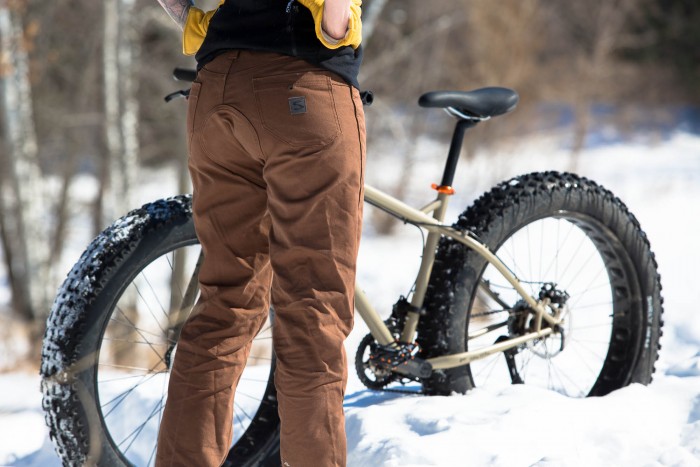 Rear, waist down view of a person wearing Surly biking pants, standing in snow, with a fat bike behind them