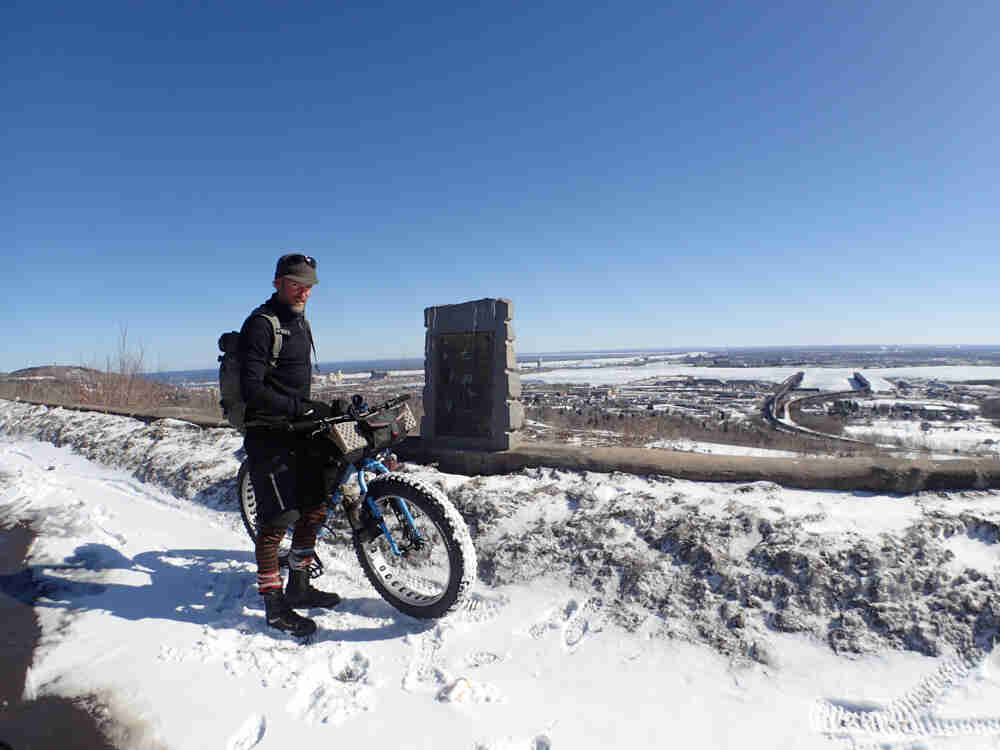 Front view of a cyclist, standing on a snow covered trail with fat bike, on top of a ledge that overlooks a city below