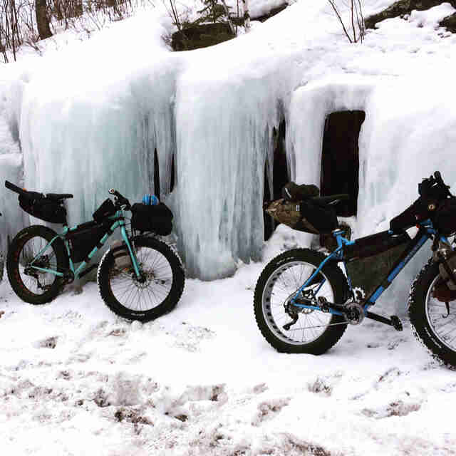 Right side view of 2 Surly fat bikes loaded with packs, parked in the snow next to a wall of icicles