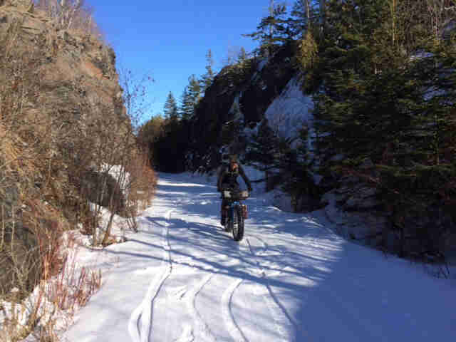 Front view of a cyclist on a fat bike, riding down a snow covered trail between rock cliffs with trees 