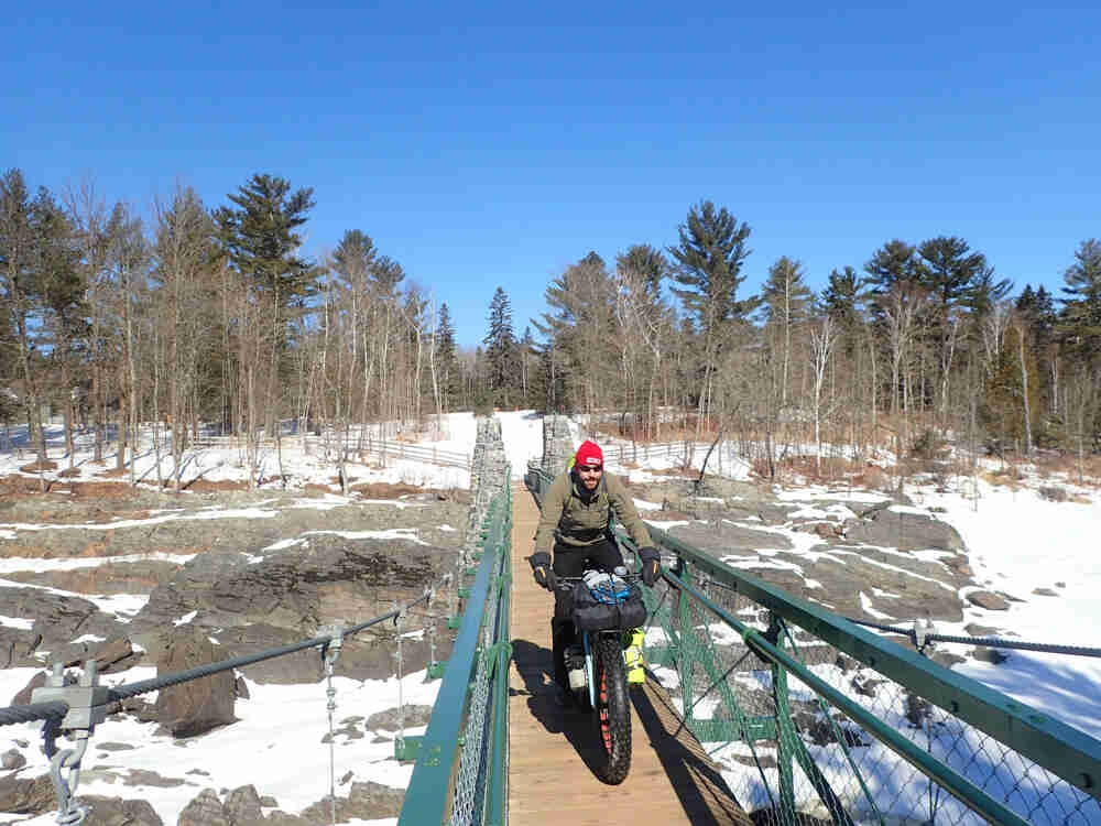Front view of a cyclist, riding a fat bike across a pedestrian bridge over a rock shoreline, with snow and trees behind