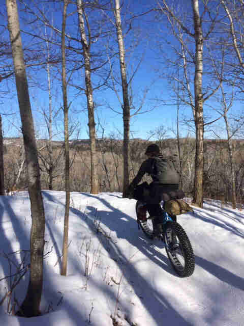 Rear view of a cyclist riding a blue fat bike with gear packs, across a snow covered trail in the woods