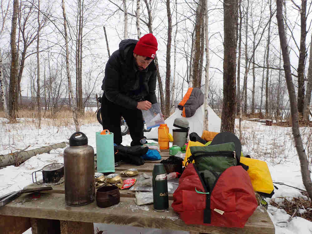 A person standing behind a table covered with gear, in a snowy campsite in the woods