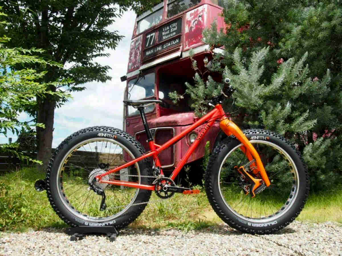Right side view of a red Surly fat bike, parked in front of a double decker bus peeking out from a tree
