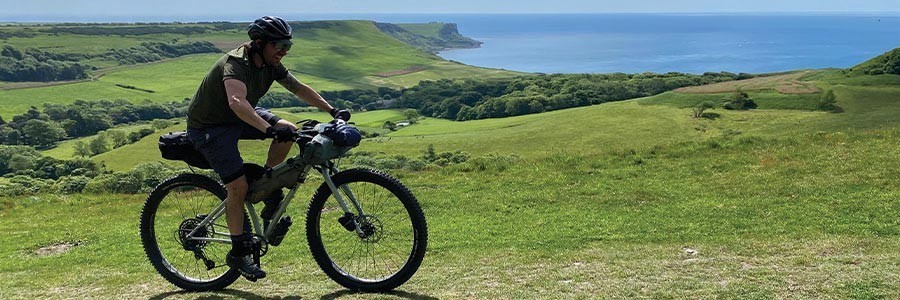 Cyclist w/ bikepacking bags riding grassy hills on sunny day with shoreline in distance
