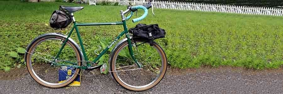 Right side view of a green Surly Pack Rat with gear packs on a sidewalk in front of a yard with a white picket fence