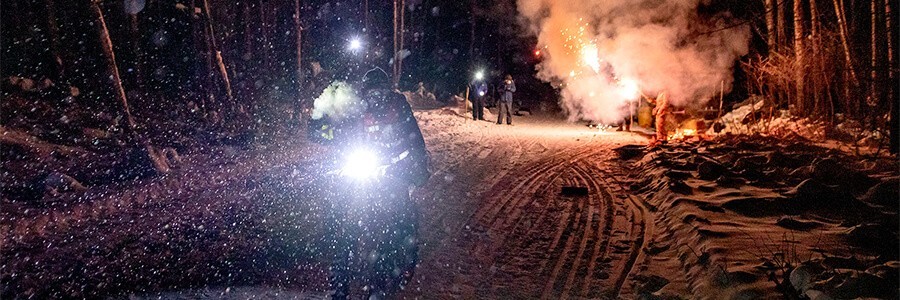 Cyclist riding a bike with a headlight, in the snow at night with fireworks exploding on the ground behind 