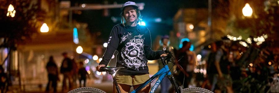 Person standing behind blue single speed Karate Monkey bike in street at night smiling