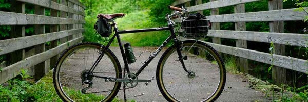 Right side view of Surly Pack Rat bike with front basket, parked on a paved trail bridge in the woods