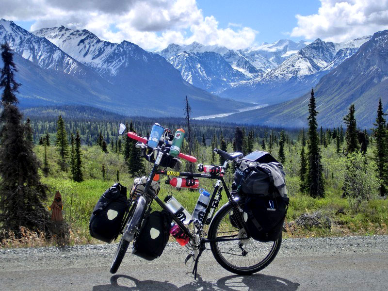 Left side view of a black bike with gear & water bottles, parked on road with a field of trees and mountains behind