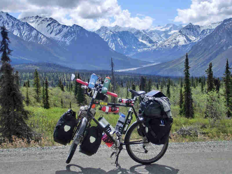 Left side view of a black bike loaded with gear, parked on a road, with a field of pines and mountains in the background