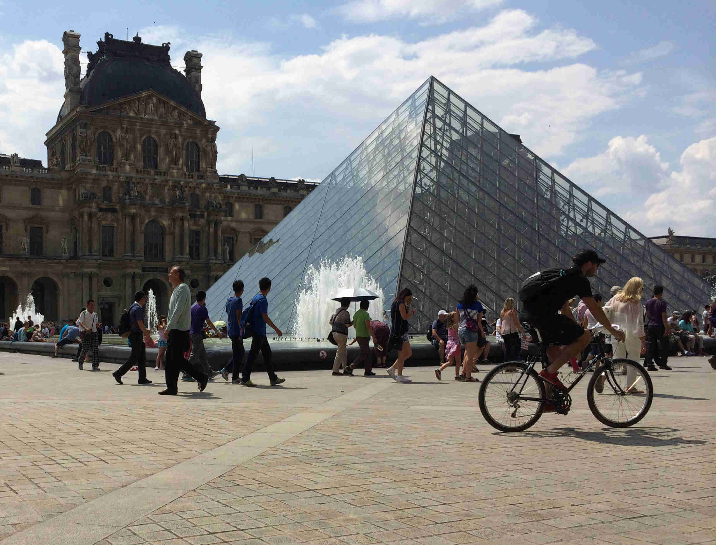 Right side view of a cyclist wearing a backpack, riding a Surly bike across the courtyard in front of the Louvre museum