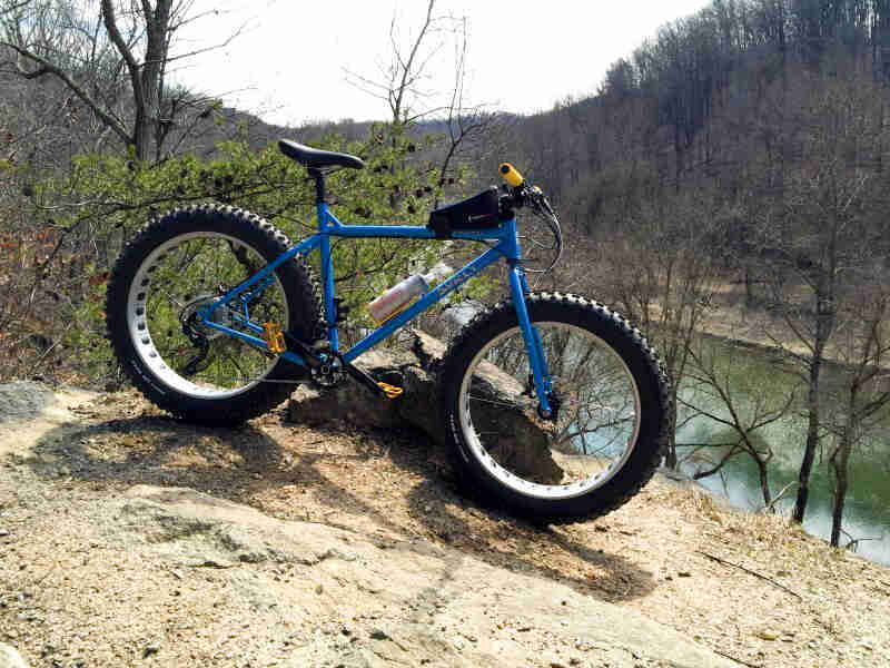 Right side view of a blue Surly fat bike, parked on a flat rock ledge above a river in the woods