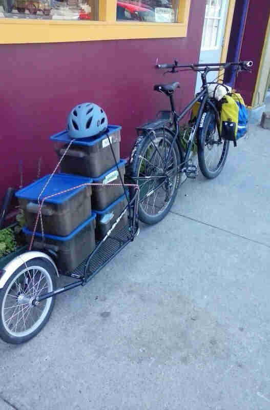 Rear, right side view of a black Surly bike with a loaded trailer of totes, leaning against a purple wall on a sidewalk