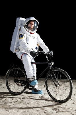 A person wearing an astronaut's suit, sitting on a bike, in front of a black background