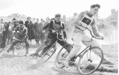 Front, right side view of cyclists, racing their bikes around a curve on a dirt race track - black & white image